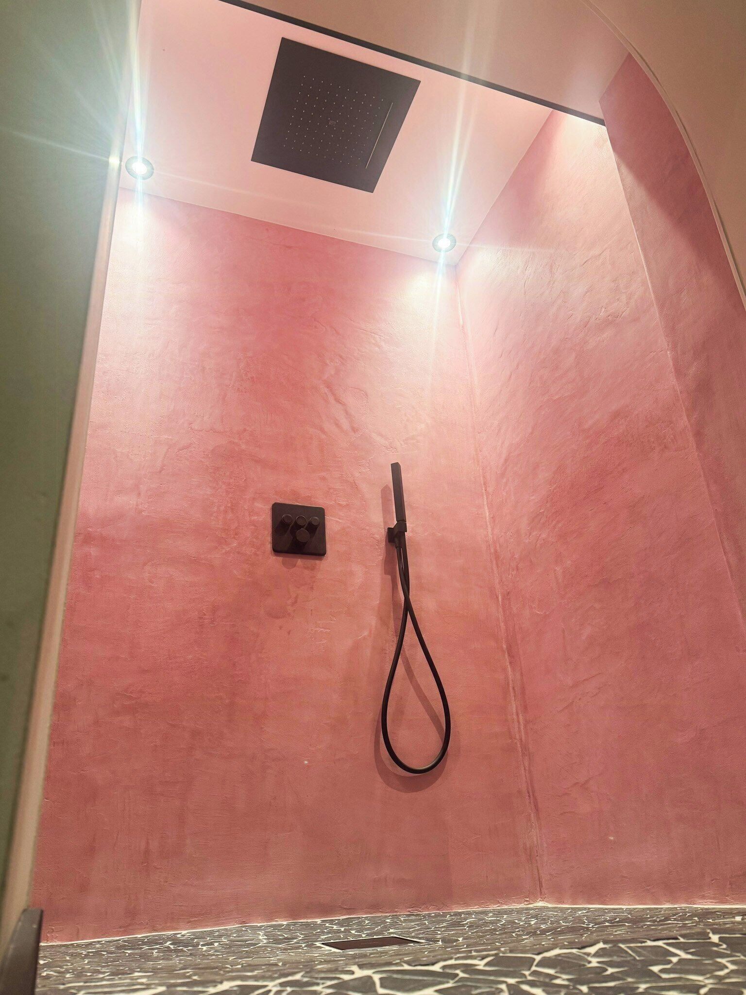 Zona de ducha con pared rosa, una lámpara en el techo y un cabezal de ducha manual.