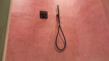 Zona de ducha con pared rosa, una lámpara en el techo y un cabezal de ducha manual.