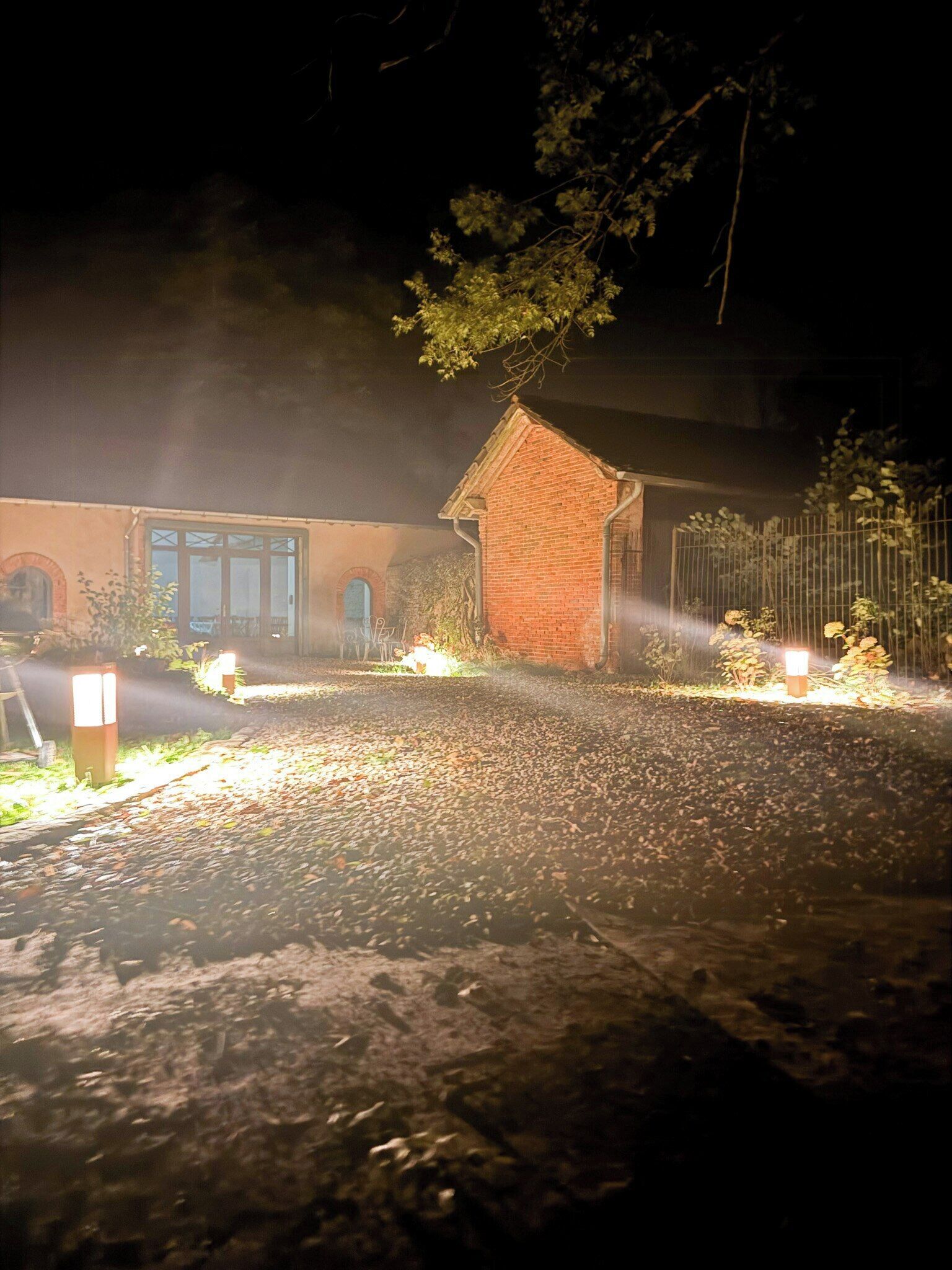 Vista nocturna de un edificio con estructura de ladrillo, una cerca de metal y un área pavimentada con hojas dispersas.