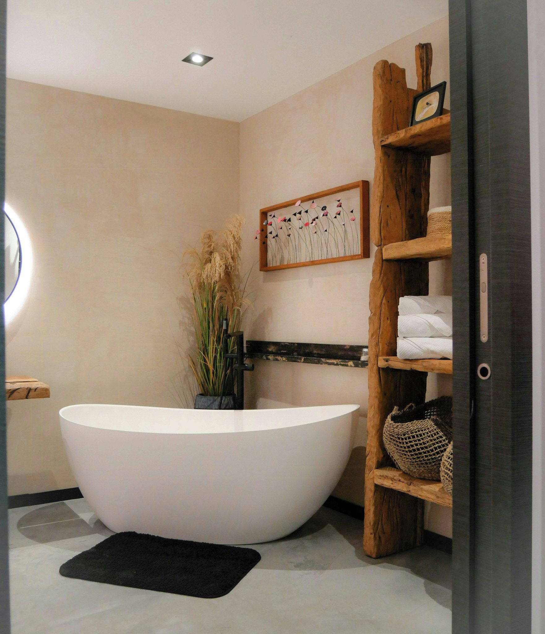 Une salle de bain moderne avec une baignoire sur pieds, une étagère en bois où sont posées des serviettes et une œuvre d’art accrochée au mur.