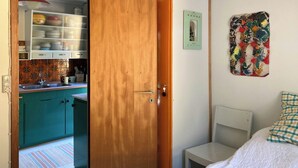 Ein Zimmer mit einem Bett, einem kleinen Nachttisch, einer Holztür, die zur Küche führt, und einem farbenfrohen, abstrakten Gemälde an der Wand.