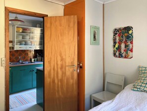 A room with a bed, a small bedside table, a wooden door leading to a kitchen, and a colorful abstract painting on the wall. - Swedish-inspired home in Odden's forest (Sjællands Odde)