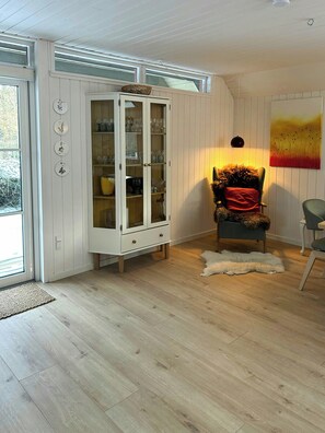 Una habitación con piso de madera, un mueble blanco con puertas de cristal, un sillón con una manta de piel y un cuadro en la pared.