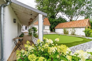 A covered outdoor seating area with a wooden deck, a lawn with a putting green, and a white building with a tiled roof. - 18th-century barn near Montreuil (Aix en Issart)
