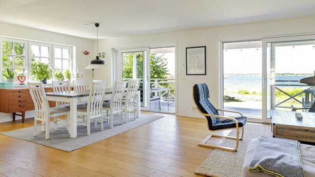 Amplia habitaci ón con piso de madera, una mesa de comedor con sillas, un sofá y vista al mar a través de amplias ventanas.
