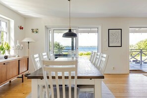 A dining area with a table and chairs, a pendant light, a cabinet, and a view of the sea through large windows.