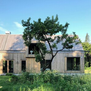A modern wooden house with a large tree in front, a visible window, and a metal roof. - Designer farm with sauna by stream in Halland (Grimeton)