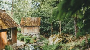 Eine rustikale Holzhütte mit Steinunterbau und einem Strohdach, umgeben von Wald und einem kleinen Bach.
