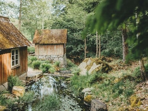 A rustic cabin with a stone foundation and a thatched roof, surrounded by a forest and a small stream. - Designer farm with sauna by stream in Halland (Grimeton)