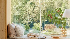 Ein gemütliches Zimmer mit einer Holzbank, Kissen, einer Lampe und Blick auf einen Wald.
