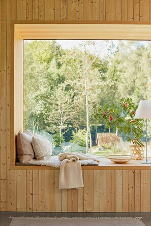 A cozy room with a wooden bench, pillows, a lamp, and a view of a forest. - Designer farm with sauna by stream in Halland (Grimeton)