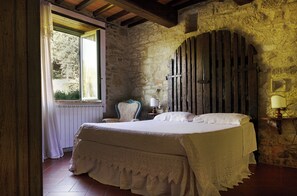A bedroom with a stone wall, a wooden door, a bed with a white bedspread, a chair, and a bedside table with a lamp. - Villa with pool in the heart of Chianti Rufina (Londa)