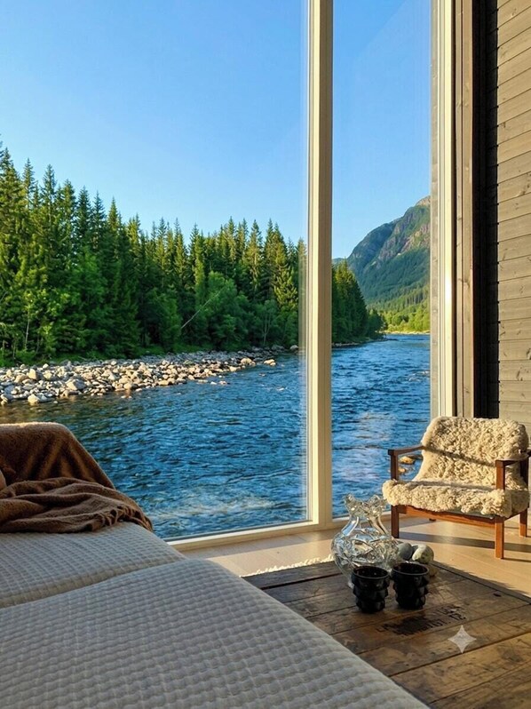 A room with a large window overlooking a river and forest, a bed, a chair, and a small table with a decorative item.