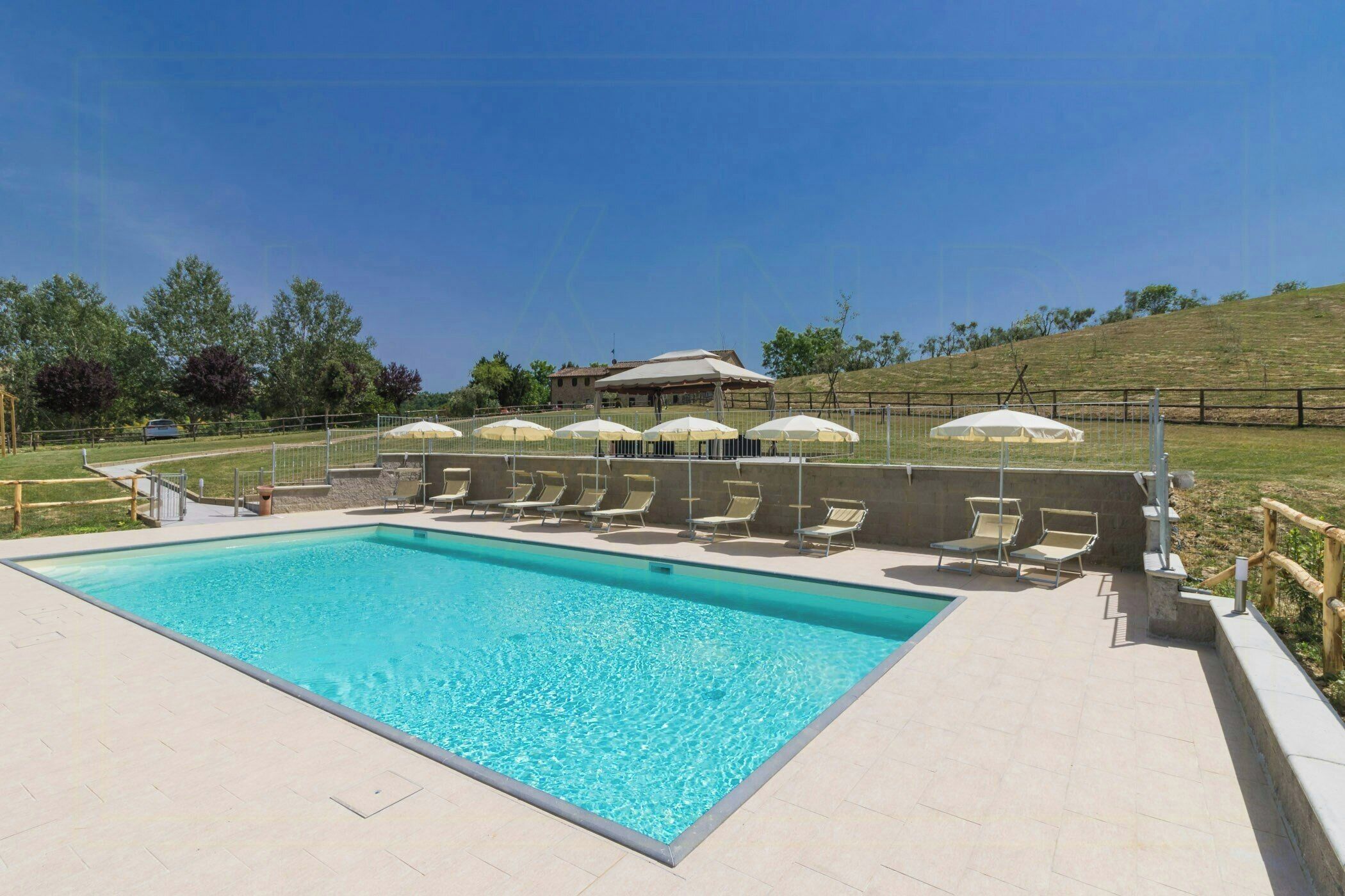 Une piscine rectangulaire à l’eau d’un bleu limpide, entourée d’une terrasse carrelée. Des chaises longues et des parasols sont disposés le long du bord de la piscine.