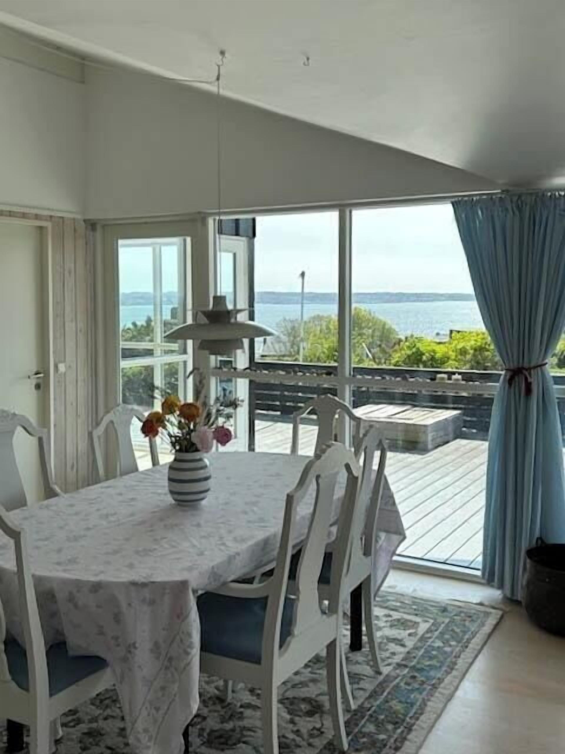 Un espace repas comprenant une table, des chaises, un vase avec des fleurs et une vue sur la mer grâce à de grandes fenêtres.