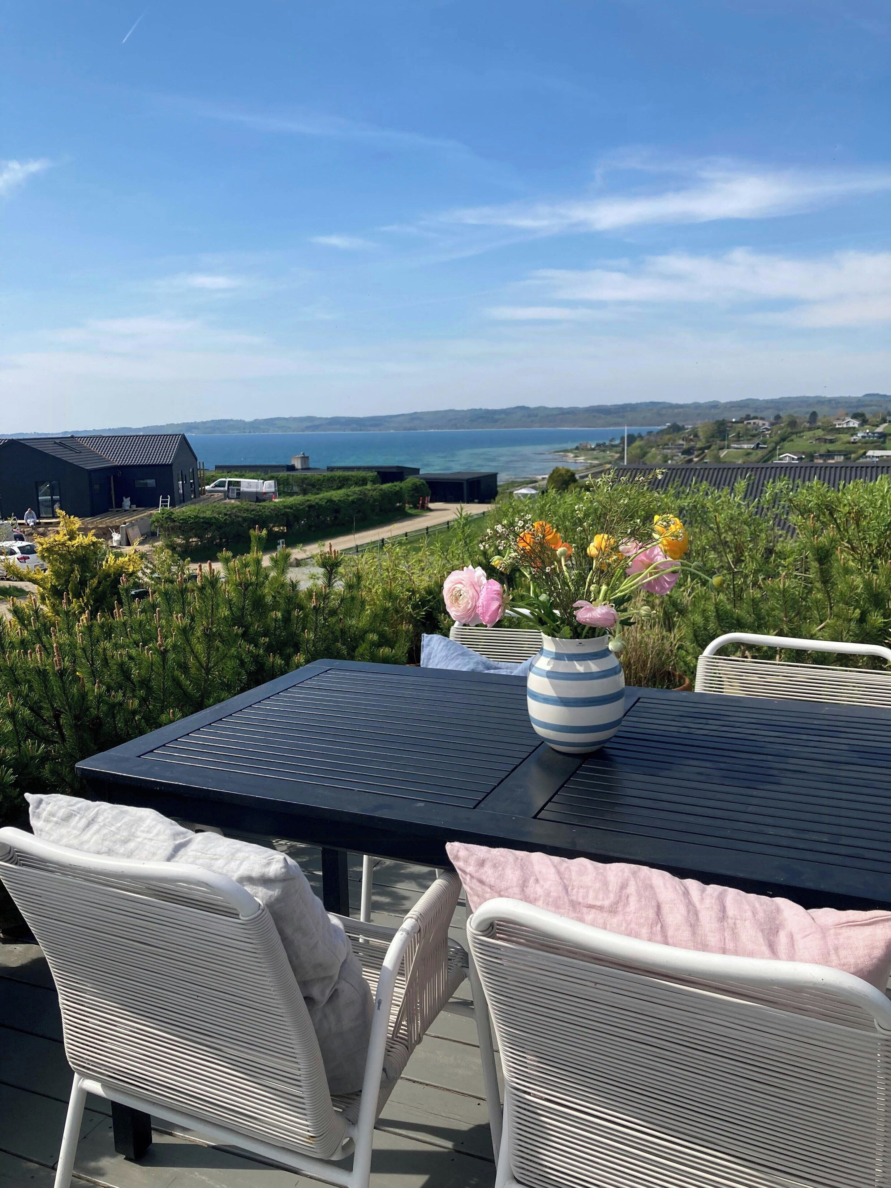 Un patio con una mesa y sillas, un florero con flores y vista al mar y colinas lejanas.