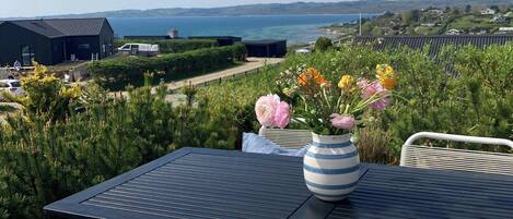 Un patio con una mesa y sillas, un florero con flores y vista al mar y colinas lejanas.