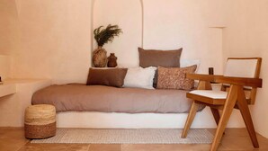 Un dormitorio acogedor con una cama, una silla de madera, un banquillo redondo pequeño y un estante en la pared.