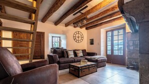 A rustic interior with exposed wooden beams, a leather sofa, a vintage trunk as a coffee table, and a stone fireplace. - Former silkworm farm from 1871 in Ardèche (Joannas)