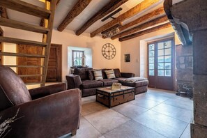 Ein rustikaler Innenraum mit freiliegenden Holzbalken, einem Ledersofa, einem alten Koffer als Couchtisch und einem Steinkamin.