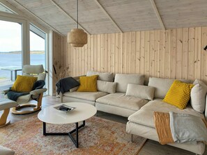 Ein gemütliches Wohnzimmer mit einer Couch, einem Sessel und einem Couchtisch, Holzwandverkleidung und Blick ins Freie.