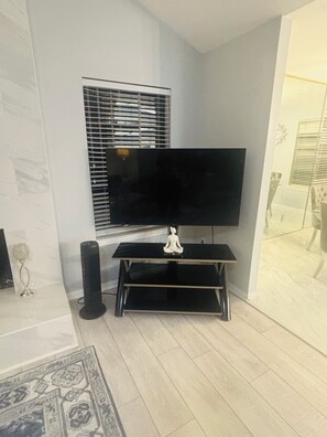 Una sala de estar con una televisión de pantalla plana sobre un mueble, una ventana con persianas, una estatua decorativa y una alfombra estampada.