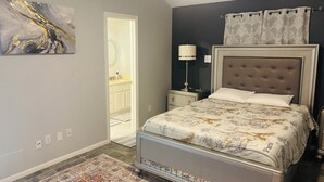 Een slaapkamer met een bed, een nachtkastje, een lamp, een decoratief wandobject en een deur naar een badkamer.