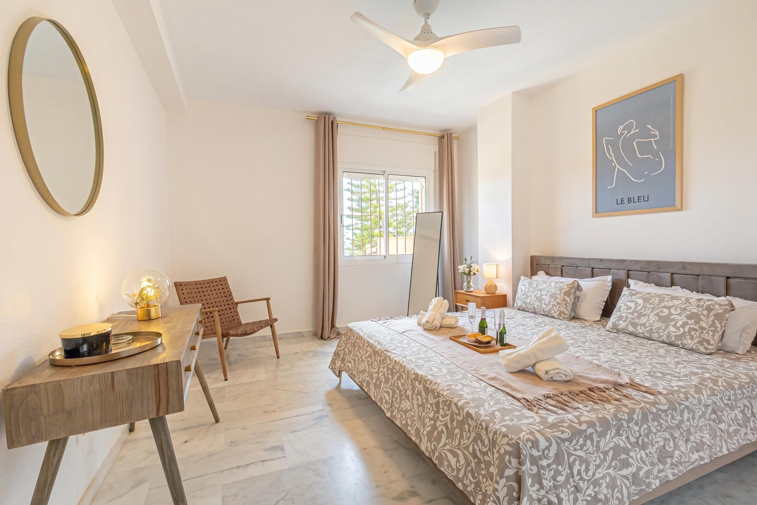 Une chambre à coucher comprenant un lit, un bureau, une chaise, un ventilateur de plafond et une fenêtre avec des rideaux.