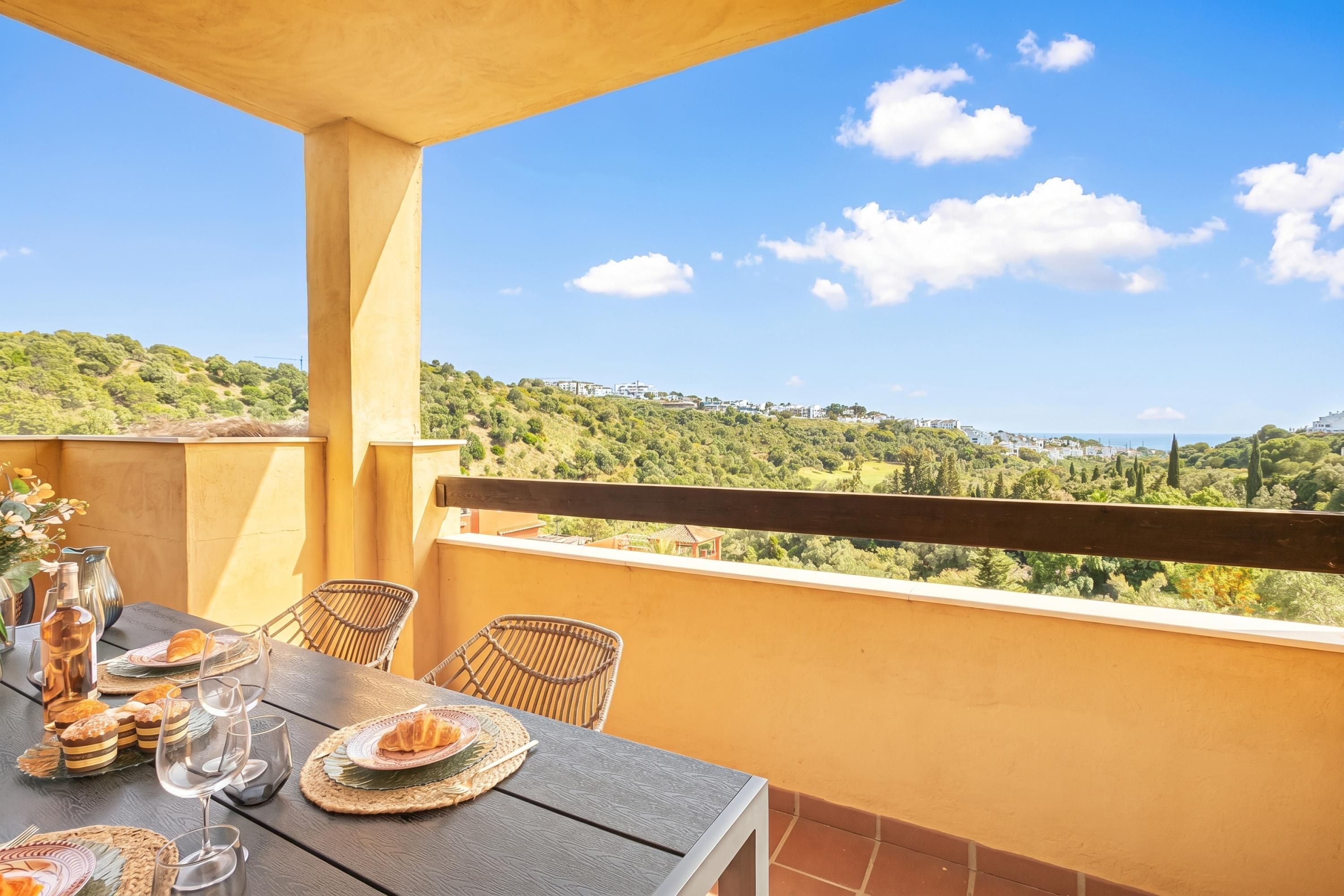 Un balcón con mesa lista para comer, sillas de mimbre y vista a un paisaje verde.