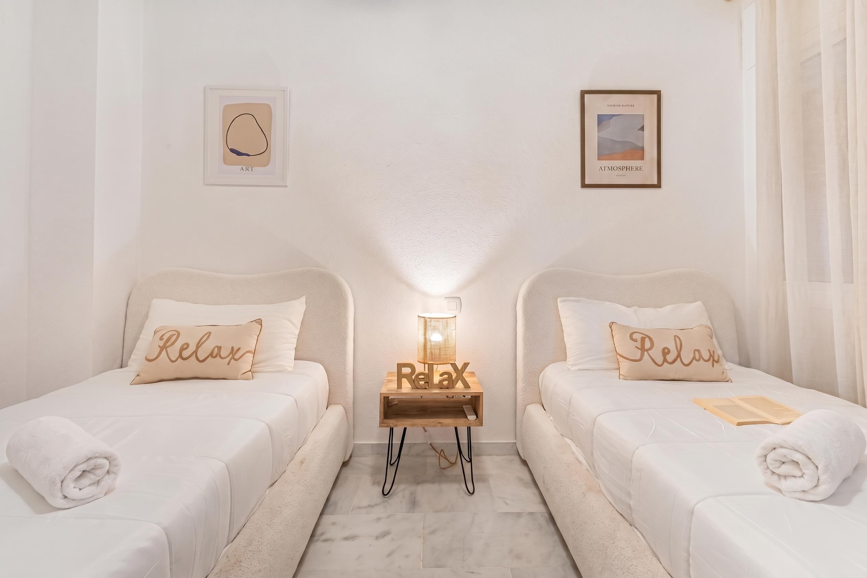 A hotel room with two beds, each with a pillow labeled 'Relax,' a bedside table with a lamp, and two framed pictures on the wall.