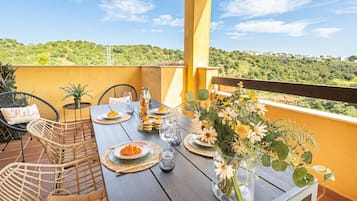 Um terraço com uma mesa posta para refeições, cadeiras de vime, um vaso com flores e vista para uma paisagem verde.