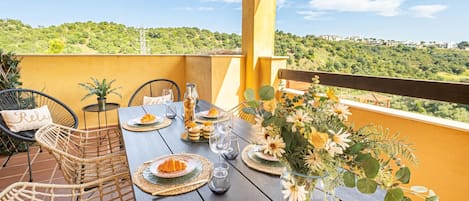 Une terrasse avec une table dressée pour le repas, des chaises en osier, un vase rempli de fleurs et une vue sur un paysage verdoyant.