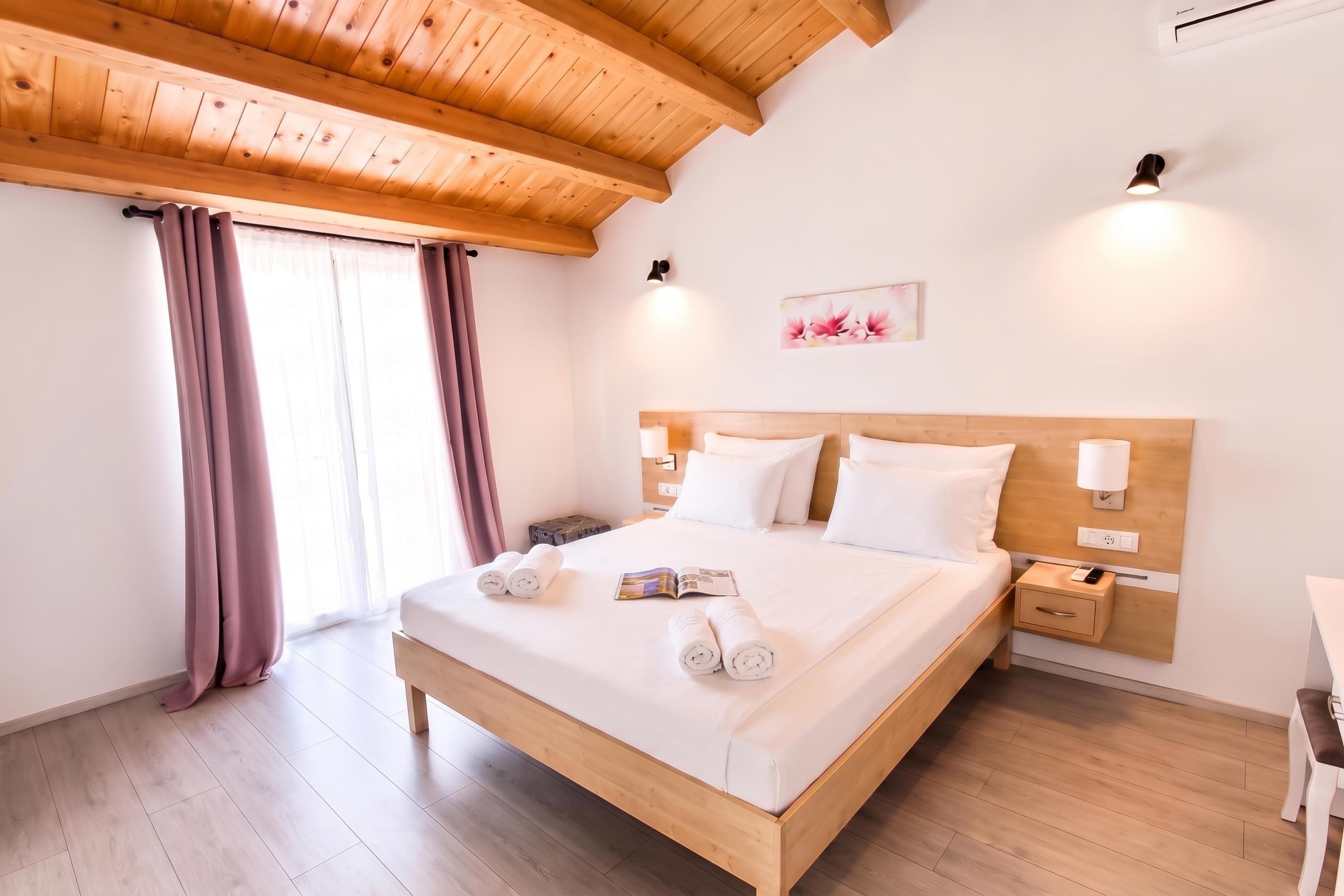 Quarto de hotel com teto de madeira, cama com roupa de cama branca, mesa de cabeceira e luminária fixada na parede.