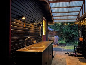 A wooden cabin interior with a kitchen area, including a sink and cabinets, and a covered outdoor deck with string lights.