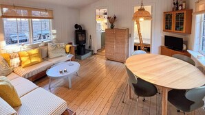 A cozy living room with a sofa, a round coffee table, a dining table with chairs, a fireplace, and a kitchen area. - Cottage by the plantation in Rørvig (Nykøbing Sjælland)