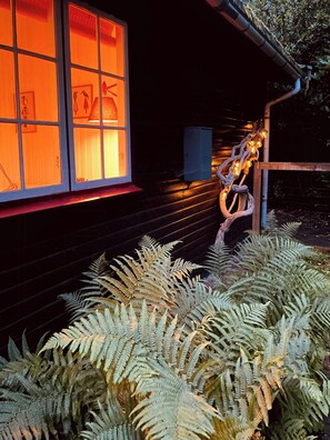 A building with a window displaying artwork, a tree wrapped in lights, and ferns in the foreground.