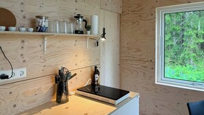 A wooden interior with a kitchen area, including a stove, shelves with kitchenware, and a window with a view of greenery. - Timeless home with nature at your doorstep (Arvika)