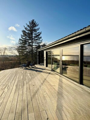 Un bâtiment moderne doté d’une terrasse, de grandes fenêtres et offrant une vue sur un arbre et un paysage.