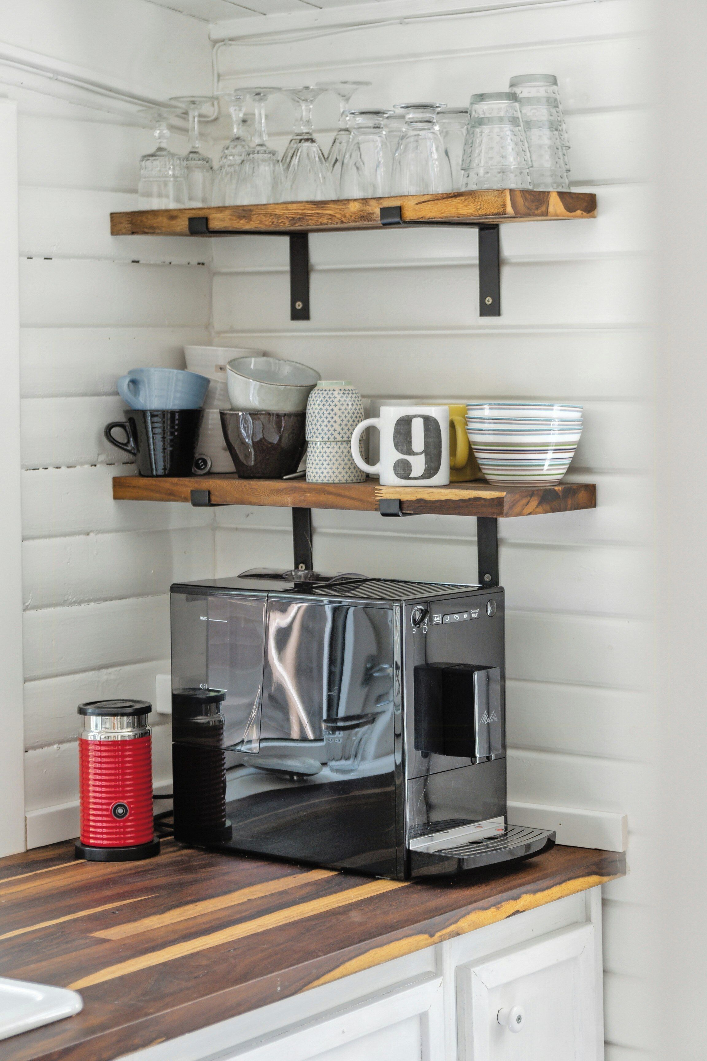 Uma cozinha com cafeteira, chaleira vermelha e uma prateleira com copos e canecas.