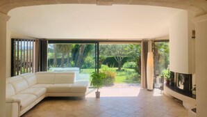 A modern living room with a white leather sofa, a flat-screen TV, and a large sliding glass door leading to a garden. - SPACIOUS VILLA, TENNIS COURT, LARGE PARK (Cuers)