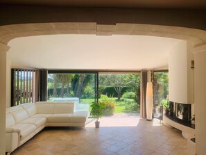 A modern living room with a white leather sofa, a flat-screen TV, and a large sliding glass door leading to a garden. - SPACIOUS VILLA, TENNIS COURT, LARGE PARK (Cuers)