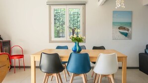 A dining area with a wooden table, four chairs, a vase with flowers, a window with a view of trees, and a painting of a beach scene. - SPACIOUS VILLA, TENNIS COURT, LARGE PARK (Cuers)