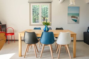 A dining area with a wooden table, four chairs, a vase with flowers, a window with a view of trees, and a painting of a beach scene. - SPACIOUS VILLA, TENNIS COURT, LARGE PARK (Cuers)