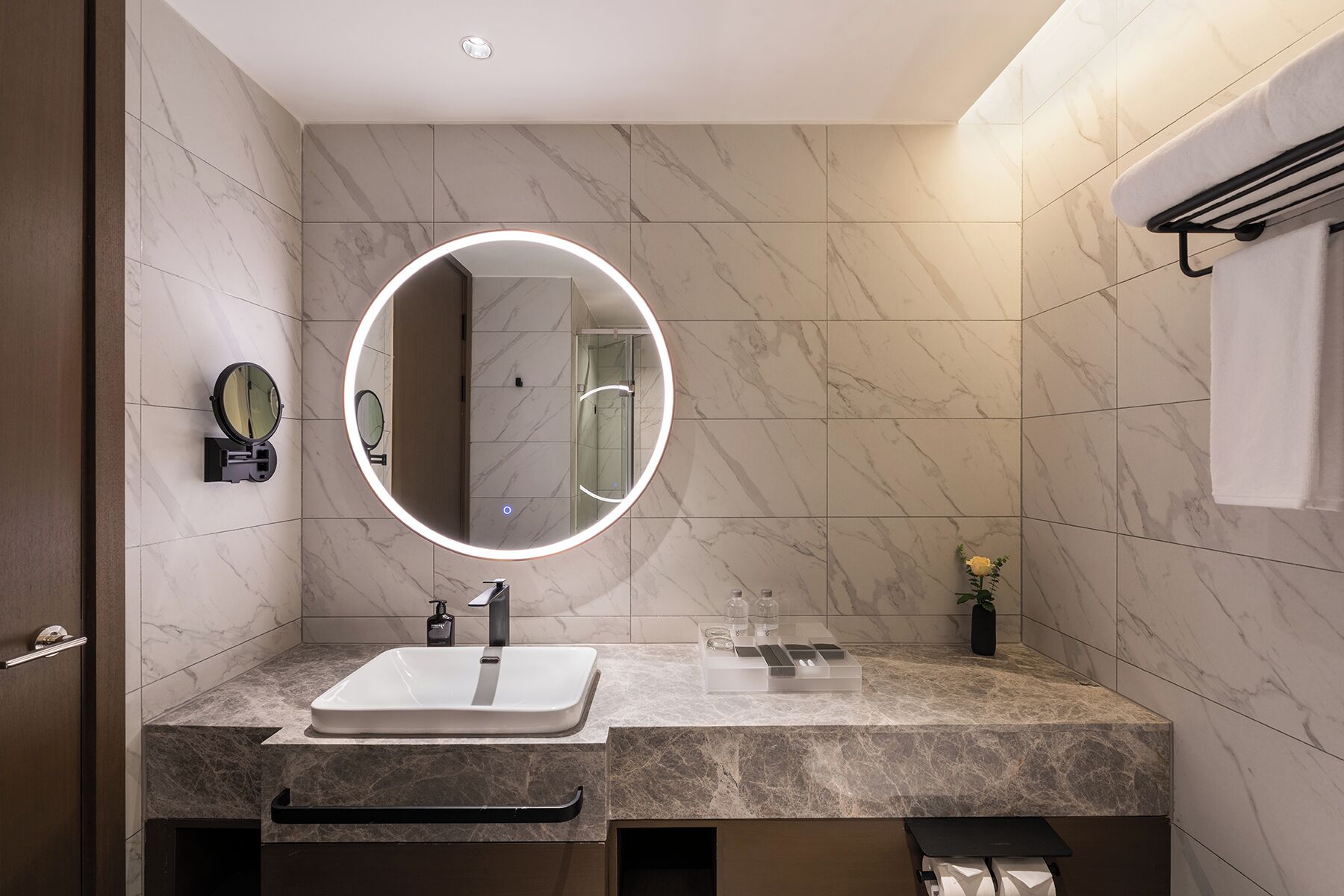 A modern bathroom with a marble sink, a round mirror with a light, and a towel rack.