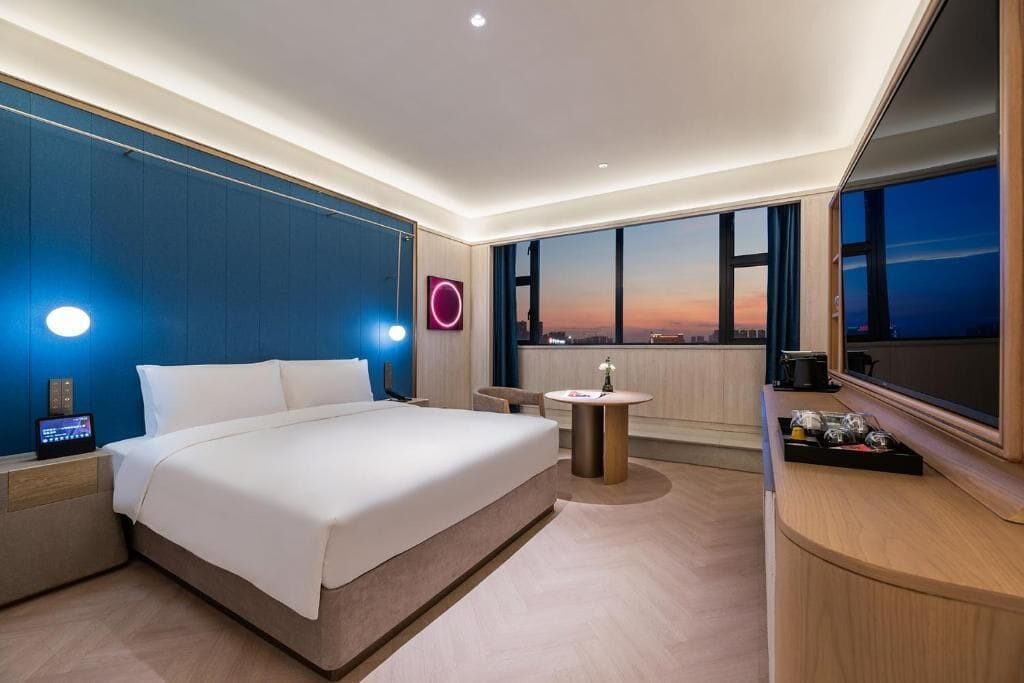 A modern hotel room with a large bed, a bedside table, a small round table, a large window with a city view, and a wall-mounted light.