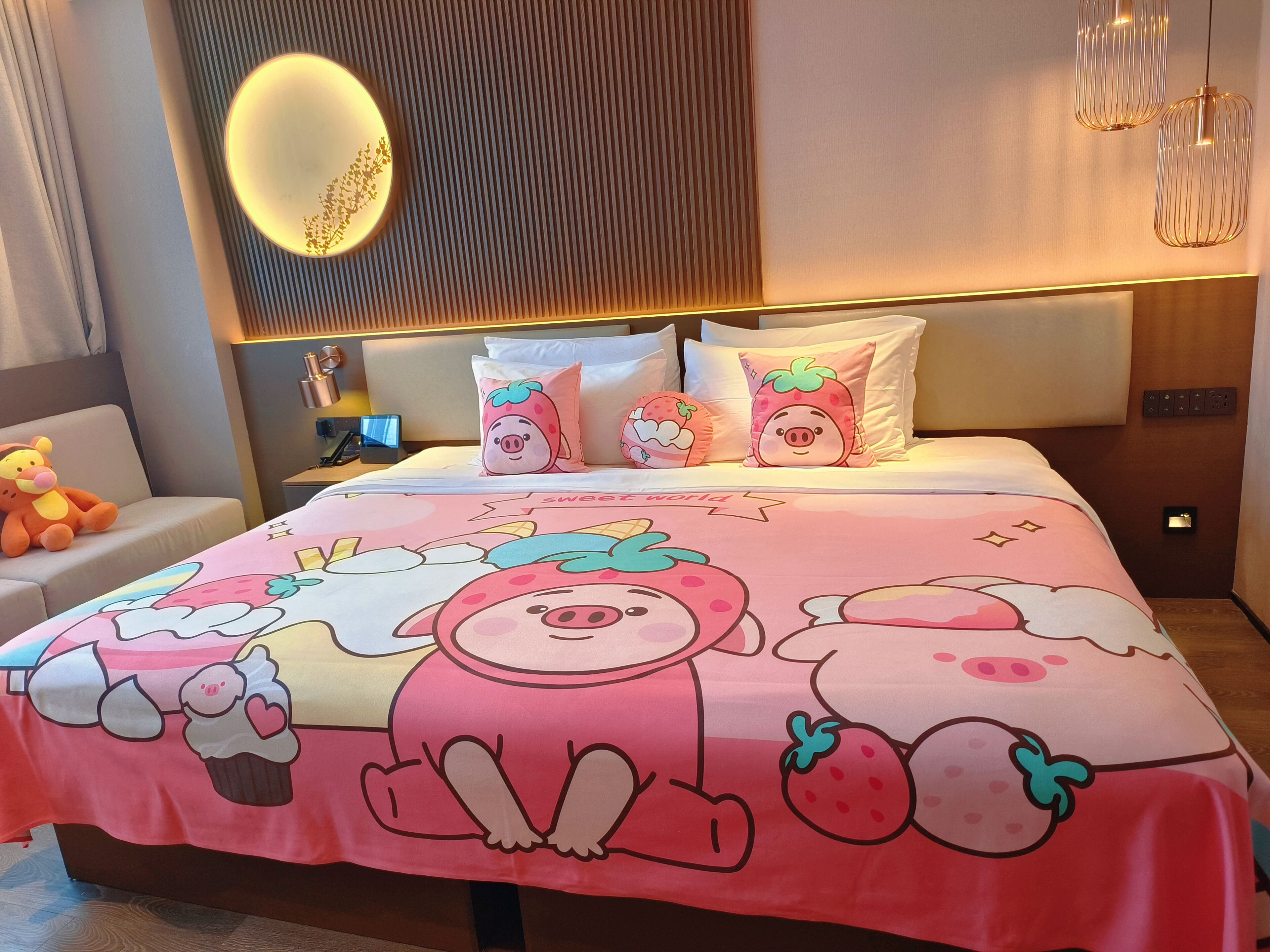 A hotel room with a bed featuring a colorful bedspread with cartoon characters, a bedside table with a lamp, a plush toy on a chair, and a modern wall design.
