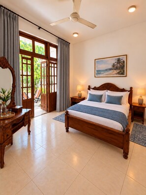 A bedroom with a wooden bed, a ceiling fan, a painting of a beach, and a view of greenery through the door. - Dimora Ceylon (Dikwella)
