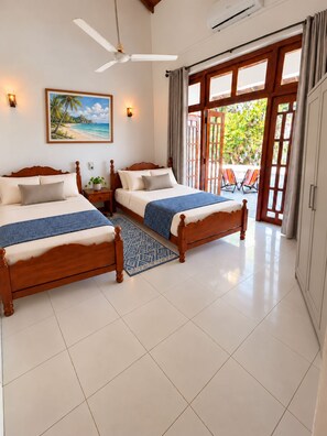 A hotel room with two wooden beds, a ceiling fan, a beach-themed painting, and a view of the outdoors through a glass door. - Dimora Ceylon (Dikwella)