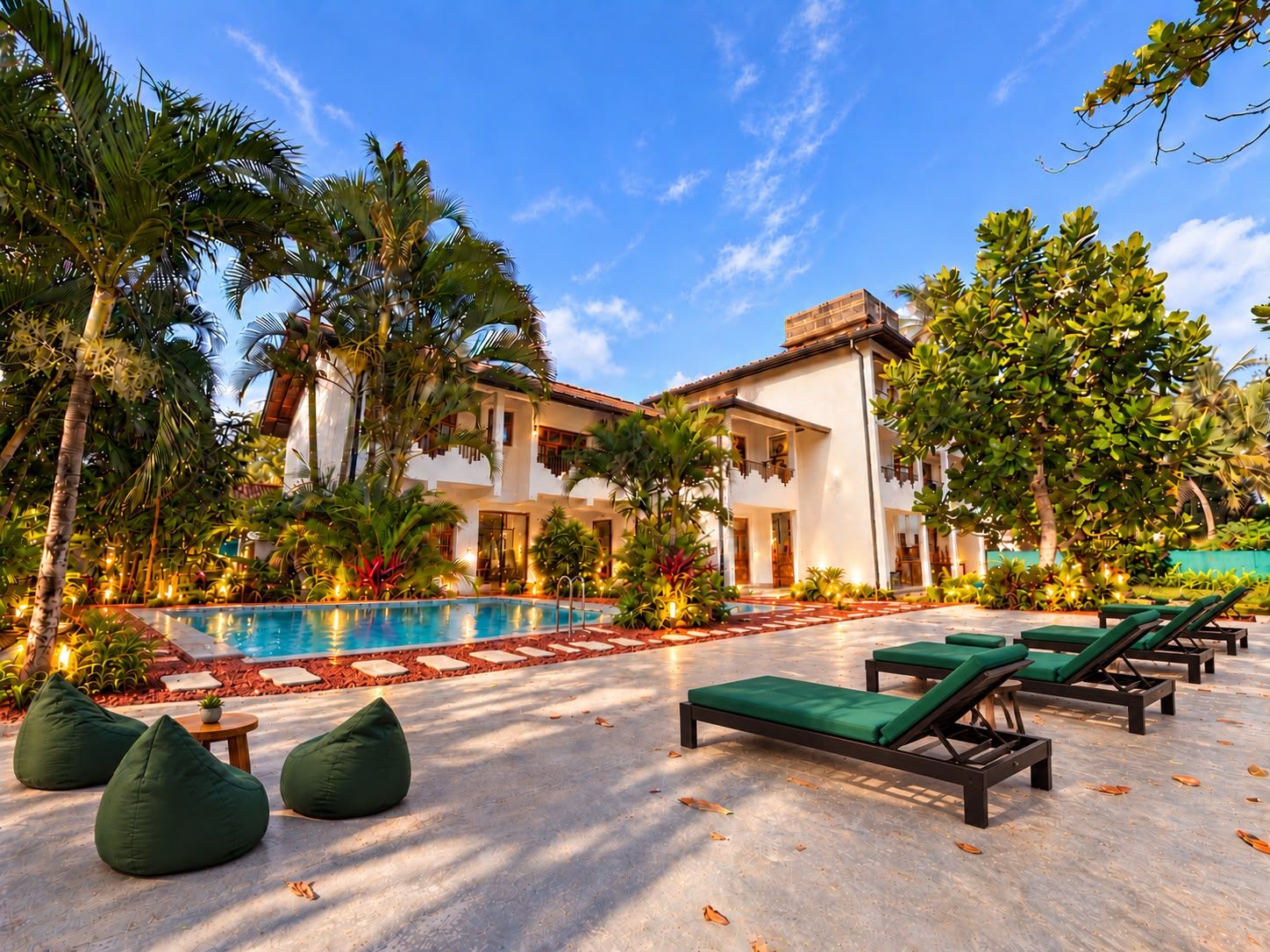 A poolside area with lounge chairs and a swimming pool, surrounded by palm trees and a building with a tiled roof.