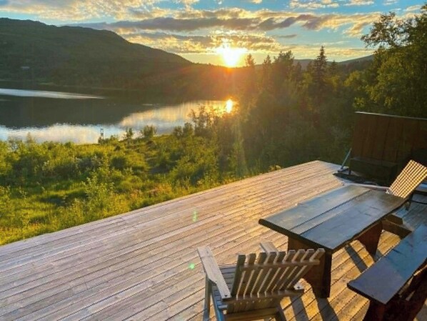 A wooden deck with a table and chairs overlooking a lake and mountains during sunset.